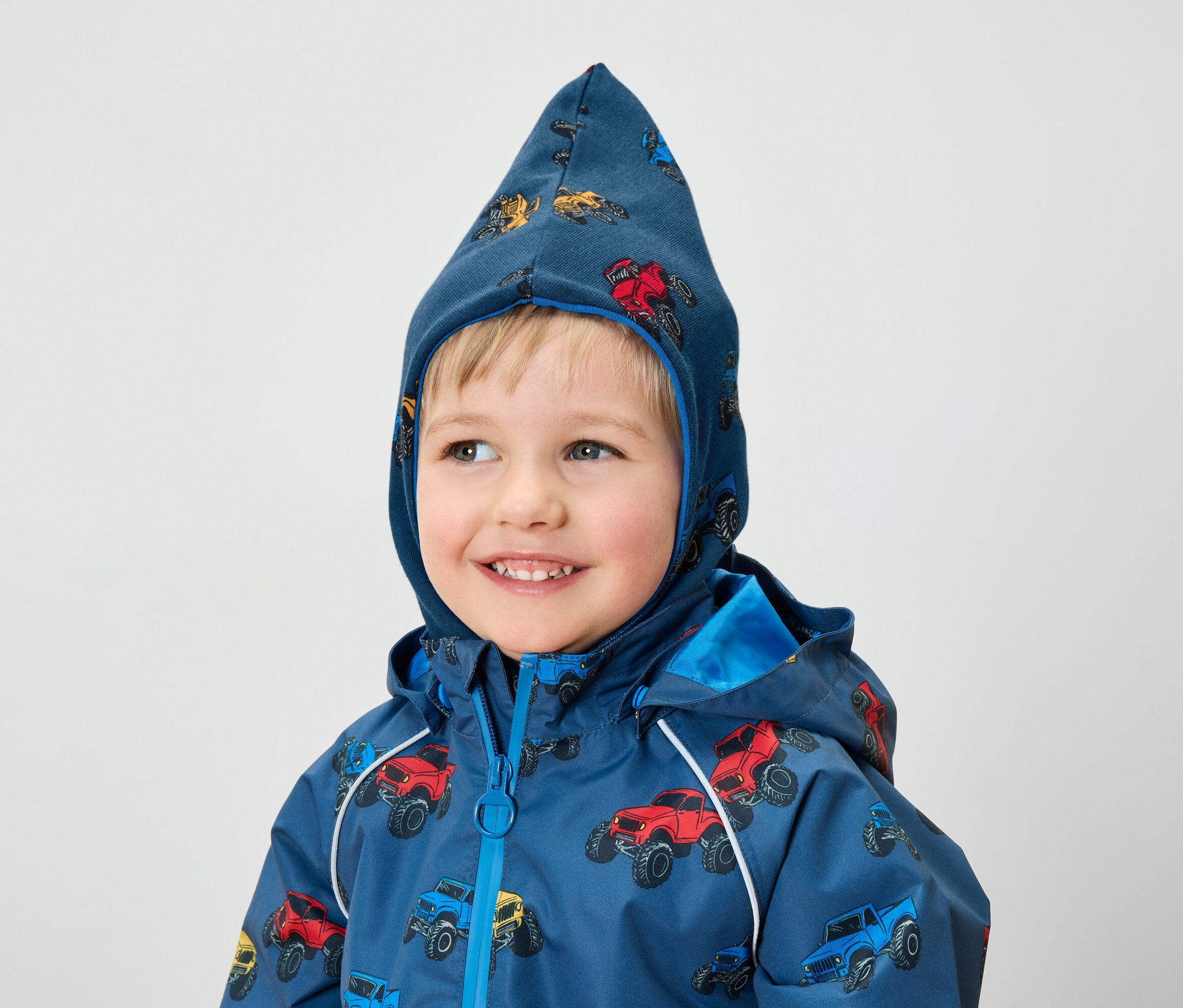 Egy mosolygó kisfiú kék kabátot és monster truck mintás kapucnit visel.