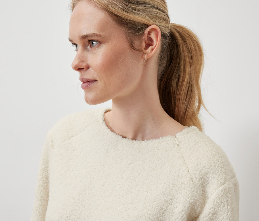 Egy szőke nő profiljának közeli képe, lófarkba kötött hajjal, krémszínű pulóvert visel.