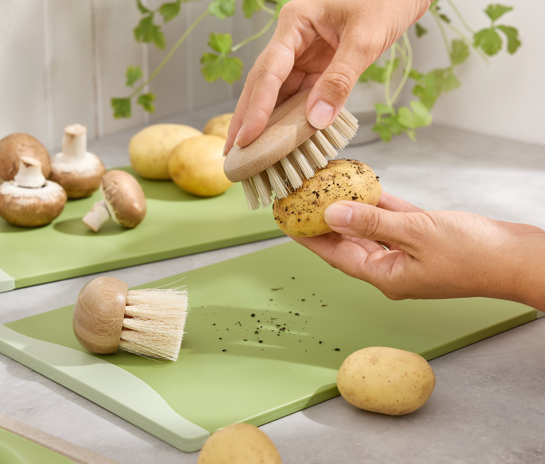 Kezek zöld vágódeszkán krumplit tisztítanak kefével. Barna csiperkegomba és krumplik a háttérben.