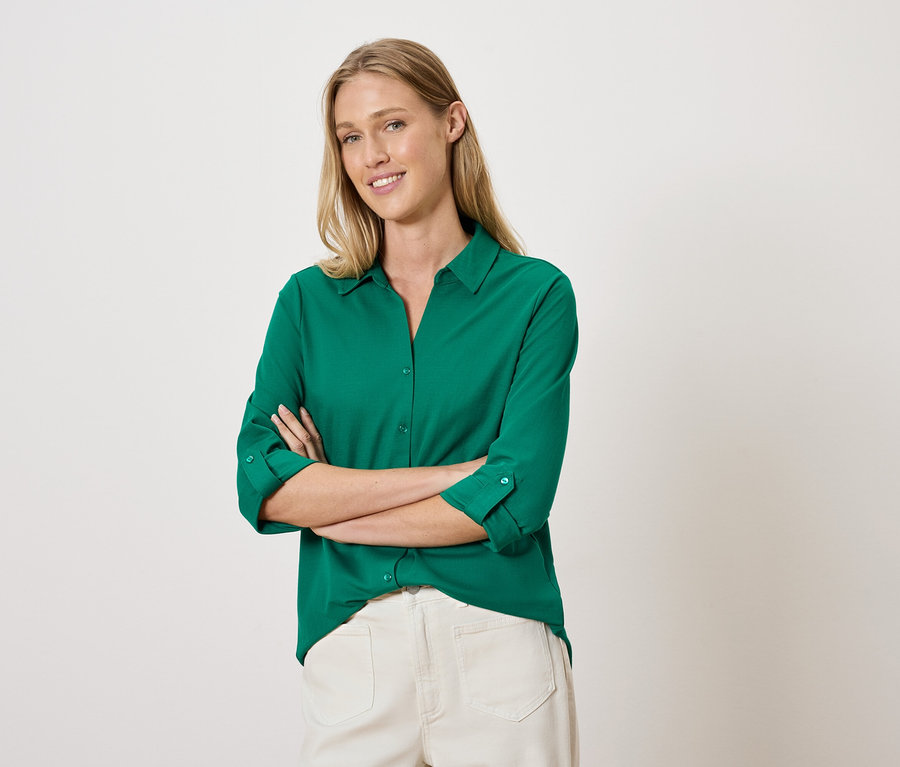 Egy nő keresztbe tett karokkal pózol egy zöld ingben és fehér nadrágban.