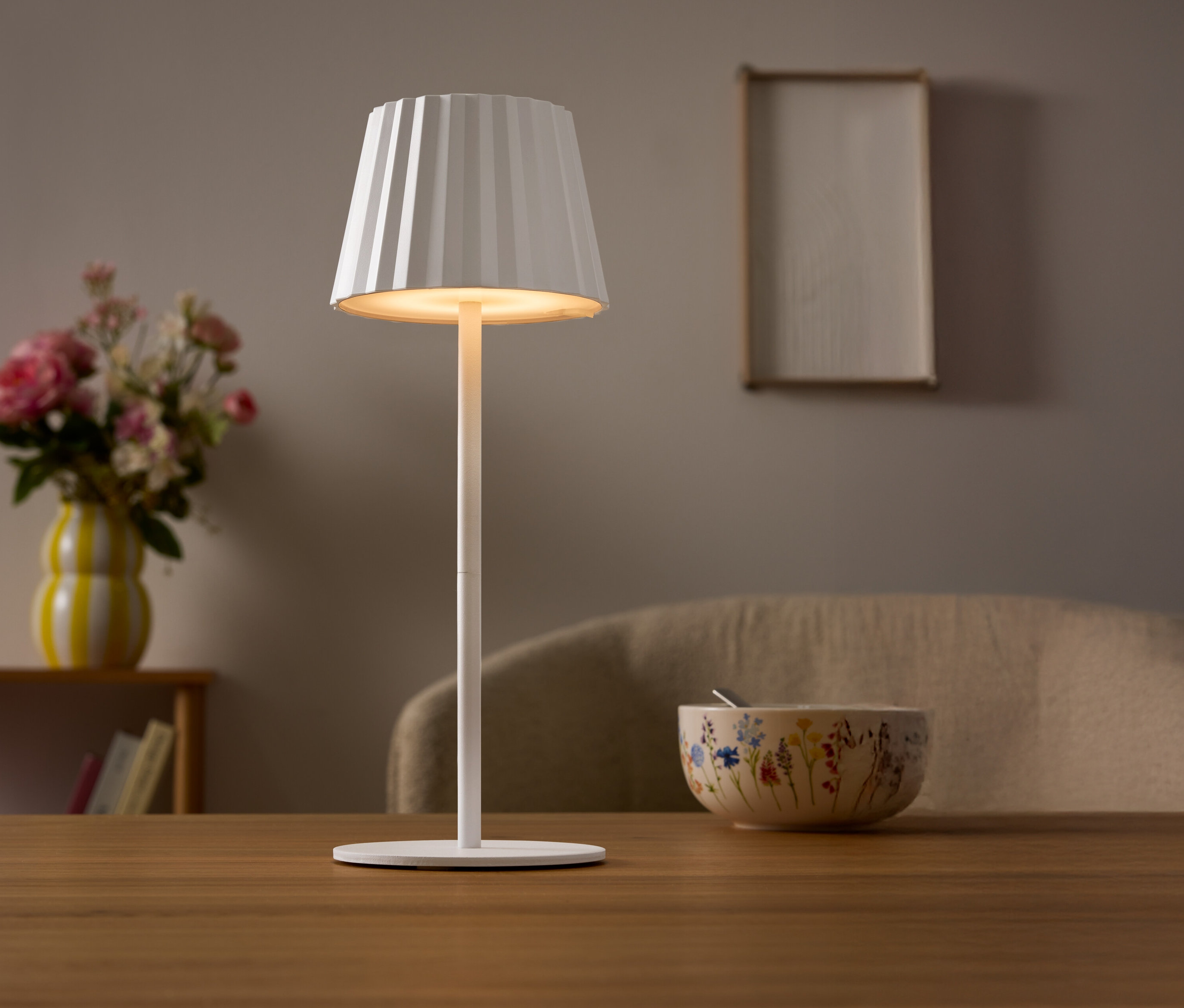 Egy asztalon egy fehér lámpa áll pliszírozott lámpabúrával. A háttérben egy virágváza és egy tál látható.