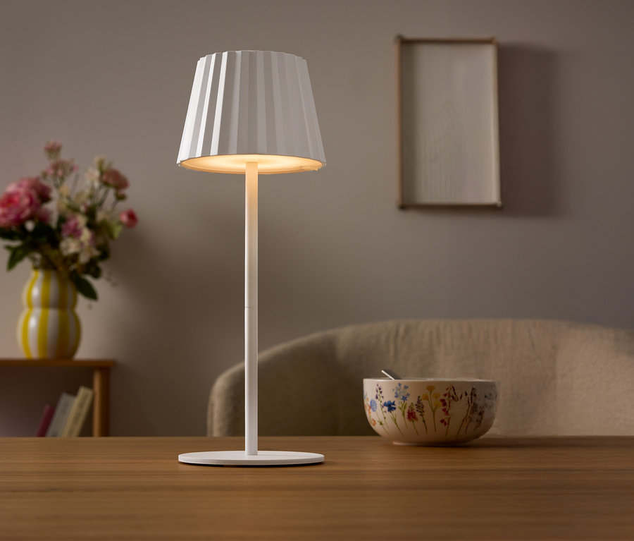 Egy asztalon egy fehér lámpa áll pliszírozott lámpabúrával. A háttérben egy virágváza és egy tál látható.