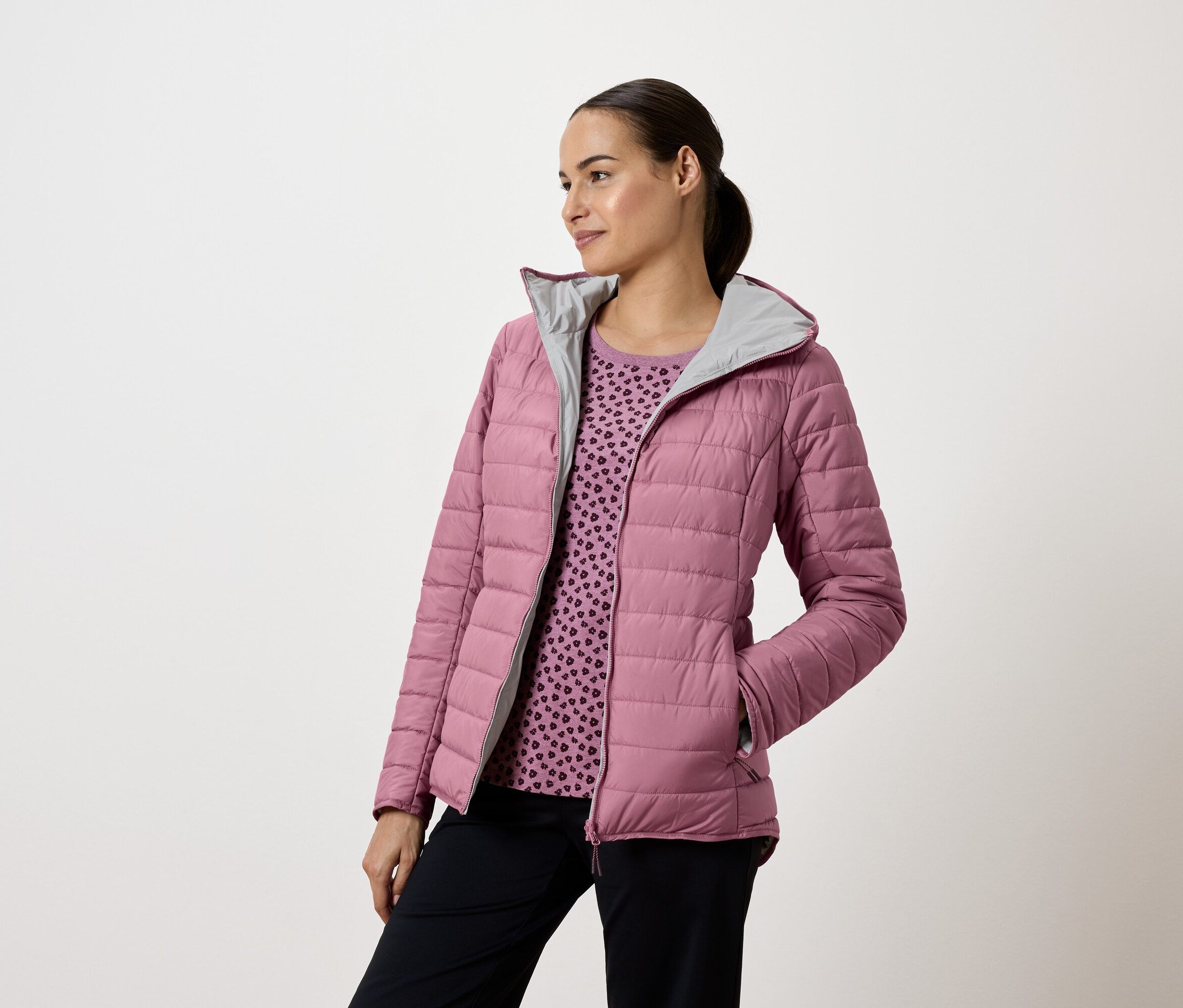 Nő rózsaszín, steppelt kabátban, kapucnival és sötétkék nadrágban, balra néz.