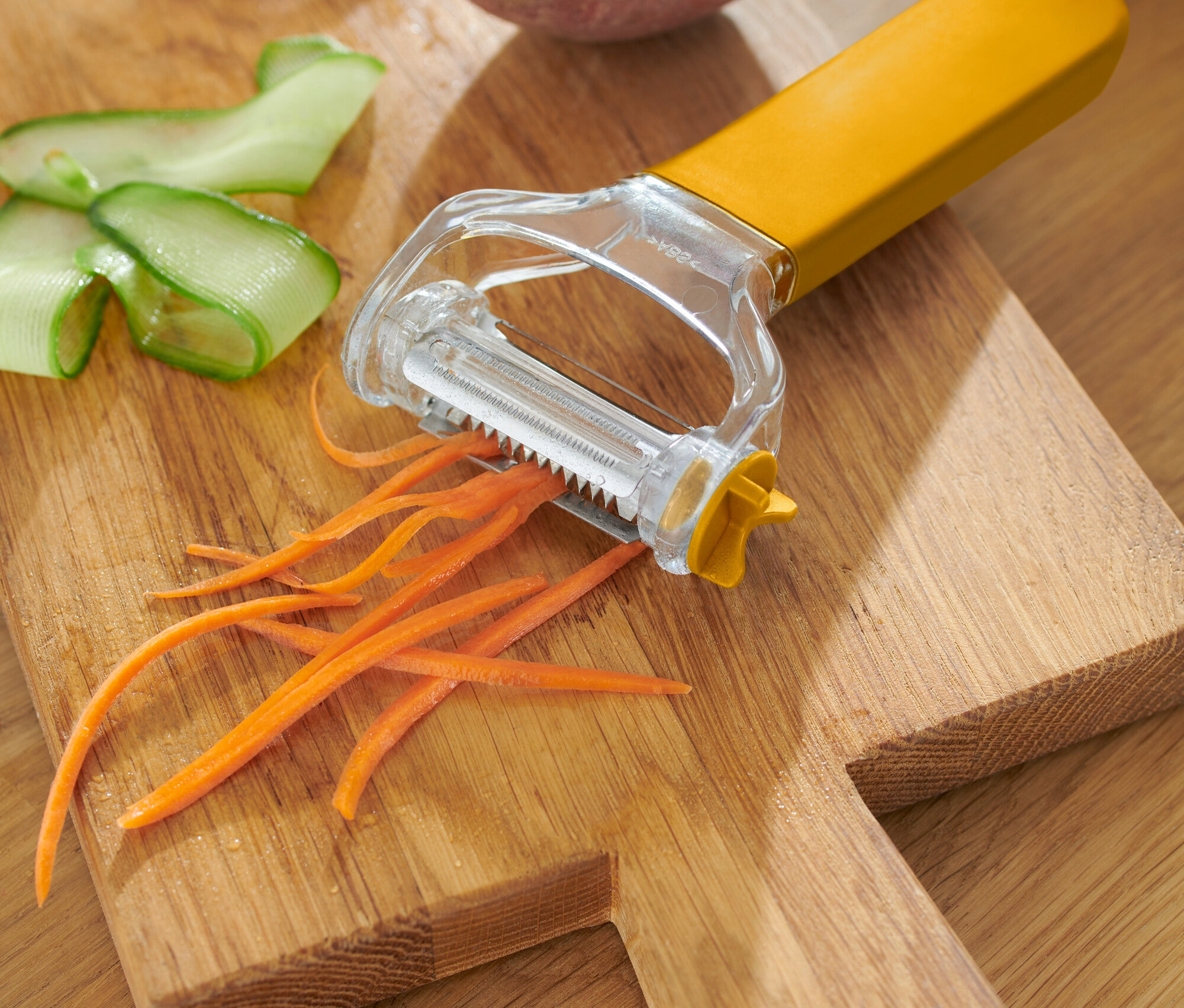 Sárga zöldséghámozó hámozott sárgarépával, uborkával és burgonyával egy fa vágódeszkán.