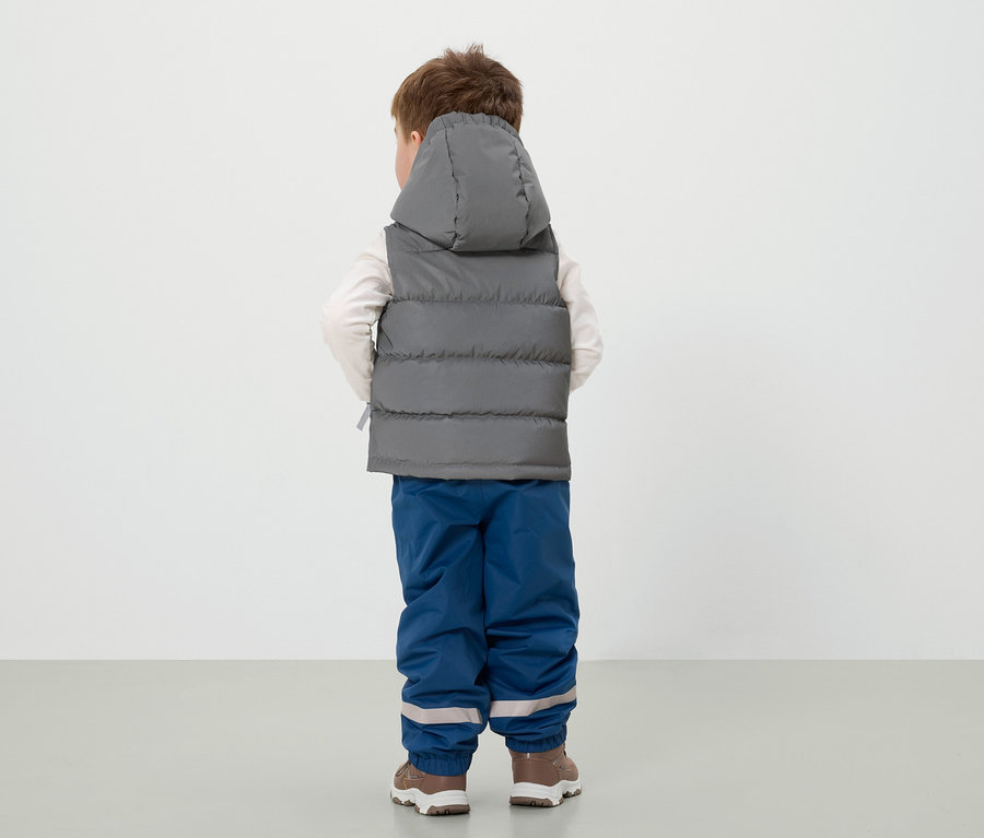 Egy fiú háttal áll, szürke kapucnis mellényt, kék nadrágot és barna cipőt visel.
