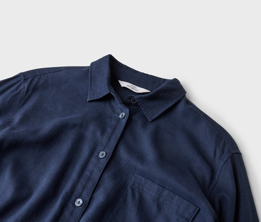 Egy sötétkék ing közeli képe, gallérral, gombokkal és egyetlen mellzsebbel semleges háttér előtt.