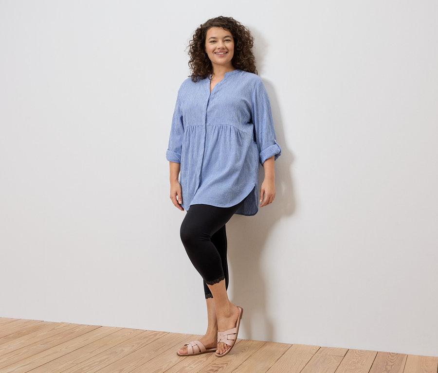 Nő áll kék ingben, csipkés fekete leggingsben és rózsaszín szandálban egy fehér fal előtt.