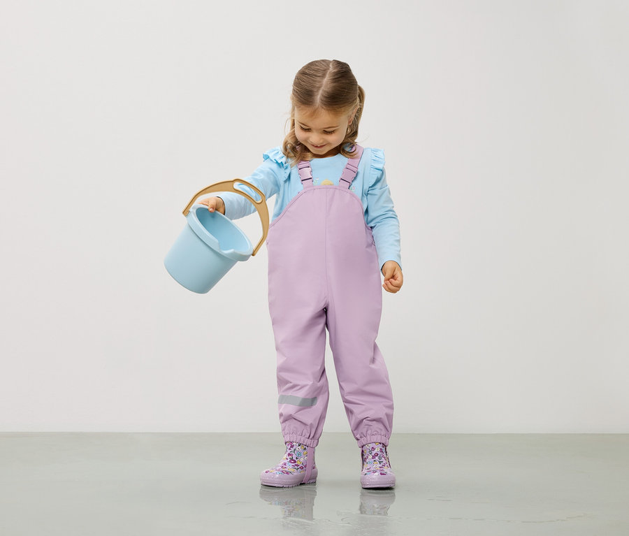 Kislány világoskék blúzban és lila overallban világoskék vödröt tart.