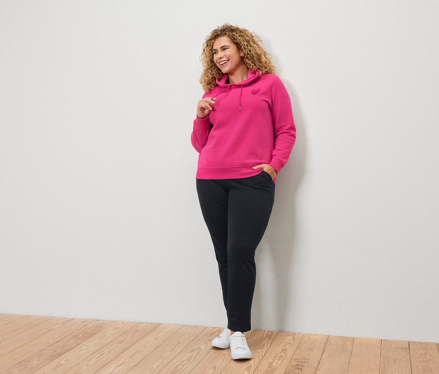 Göndör hajú nő rózsaszín kapucnis pulóverben és fekete nadrágban.