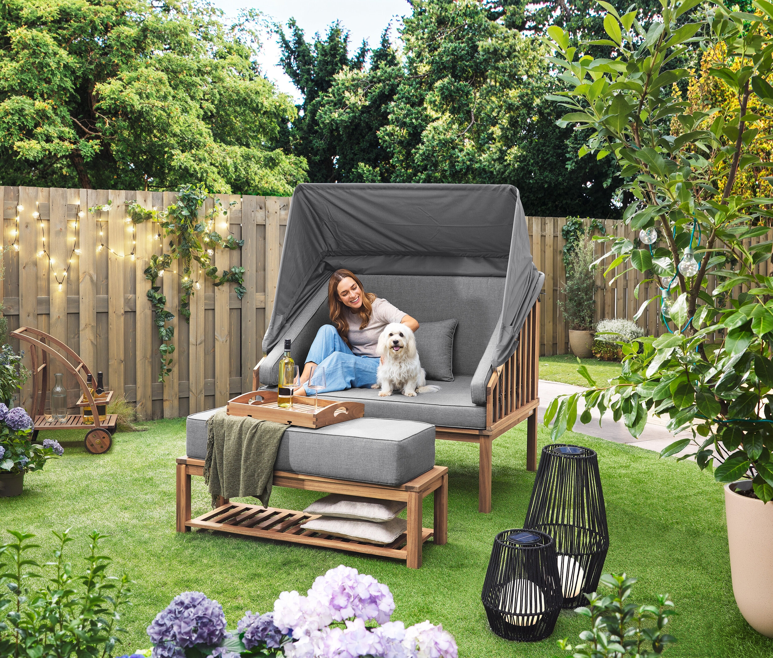 Egy nő ül egy kutyával egy Premium Lounge-Strandkorb mit Sonnenschutz-Dach nevű strandszékben. A füvön egy nagyméretű, textilpántos napelemes viharlámpa és egy kisméretű, textilszalaggal ellátott napelemes viharlámpa áll. A háttérben egy levehető tálcás bárkocsi látható. Egy LED-es kültéri fényfüzér lóg a kerítésen.