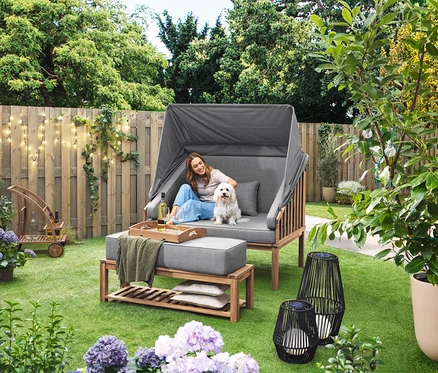 Egy nő ül egy kutyával egy Premium Lounge-Strandkorb mit Sonnenschutz-Dach nevű strandszékben. A füvön egy nagyméretű, textilpántos napelemes viharlámpa és egy kisméretű, textilszalaggal ellátott napelemes viharlámpa áll. A háttérben egy levehető tálcás bárkocsi látható. Egy LED-es kültéri fényfüzér lóg a kerítésen.