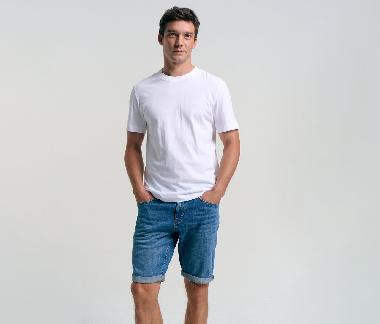 Férfi fehér pólóban és kőmosott középkék farmer Tom Tailor TTJOSH REGULAR SLIM rövidnadrágban, keze a zsebében.