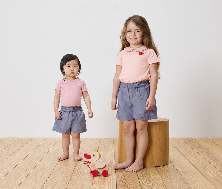 Két lány áll egy fapadlón, mindkettőjük rózsaszín felsőt és kék rövidnadrágot visel. A padlón egy fa játék található.