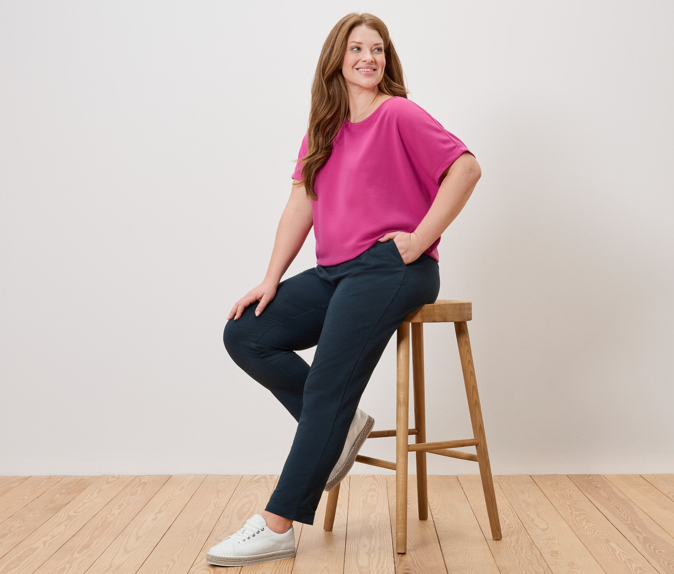 Nő ül egy széken, rózsaszín felsőt, kék nadrágot és fehér cipőt visel.