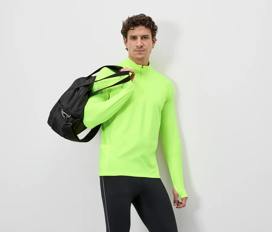 Férfi fekete sporttáskával a vállán, neon sárga hosszú ujjú felsőt és fekete leggingset visel fehér háttér előtt.