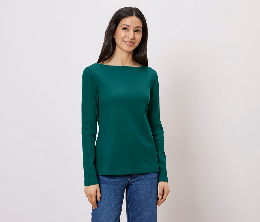 Nő visel zöld hosszú ujjú blúzt és farmert fehér háttér előtt.