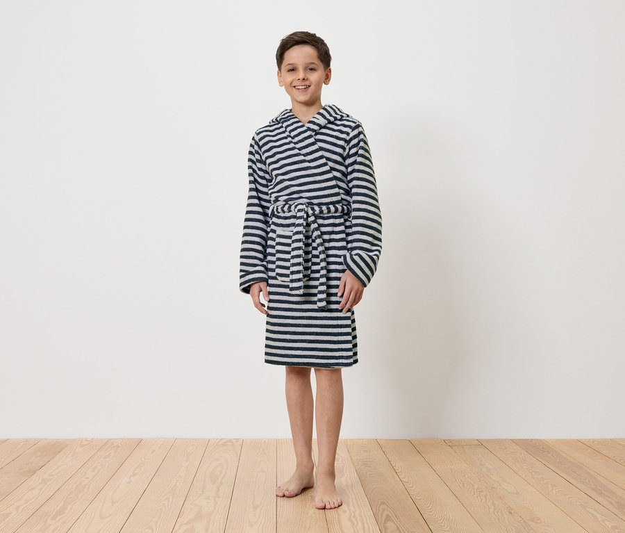 Egy fiú mezítláb áll egy fehér fal előtt, csíkos, kapucnis köntöst visel.