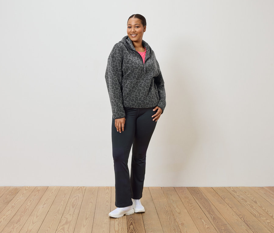 Nő áll, szürke-fekete leopárdmintás polár pulóvert, fekete nadrágot és fehér tornacipőt visel.
