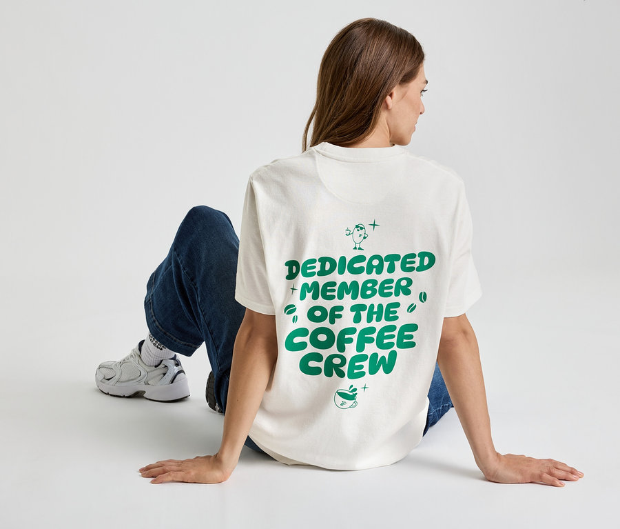 Nő ül a földön, "Dedicated Member of the Coffee Crew" feliratú pólót, farmert és tornacipőt visel.