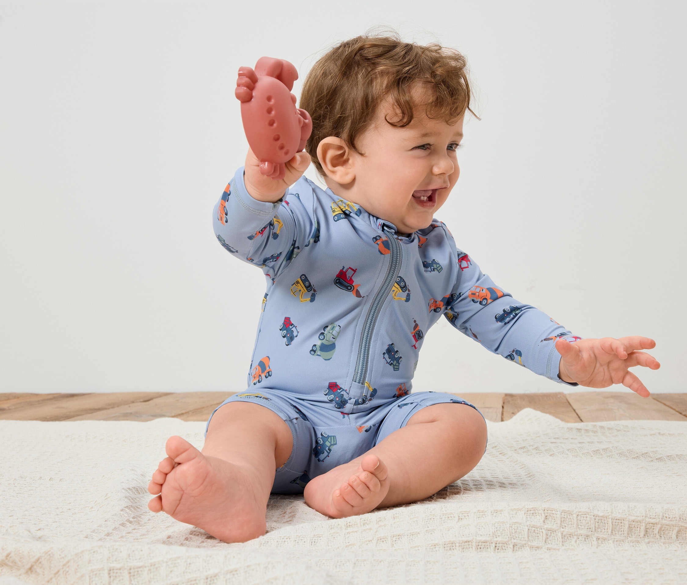 Egy mosolygó, göndör hajú csecsemő ül egy fehér takarón, és egy piros rágókát tart a kezében. A csecsemő világoskék, színes járművekkel mintázott rugdalózót visel.