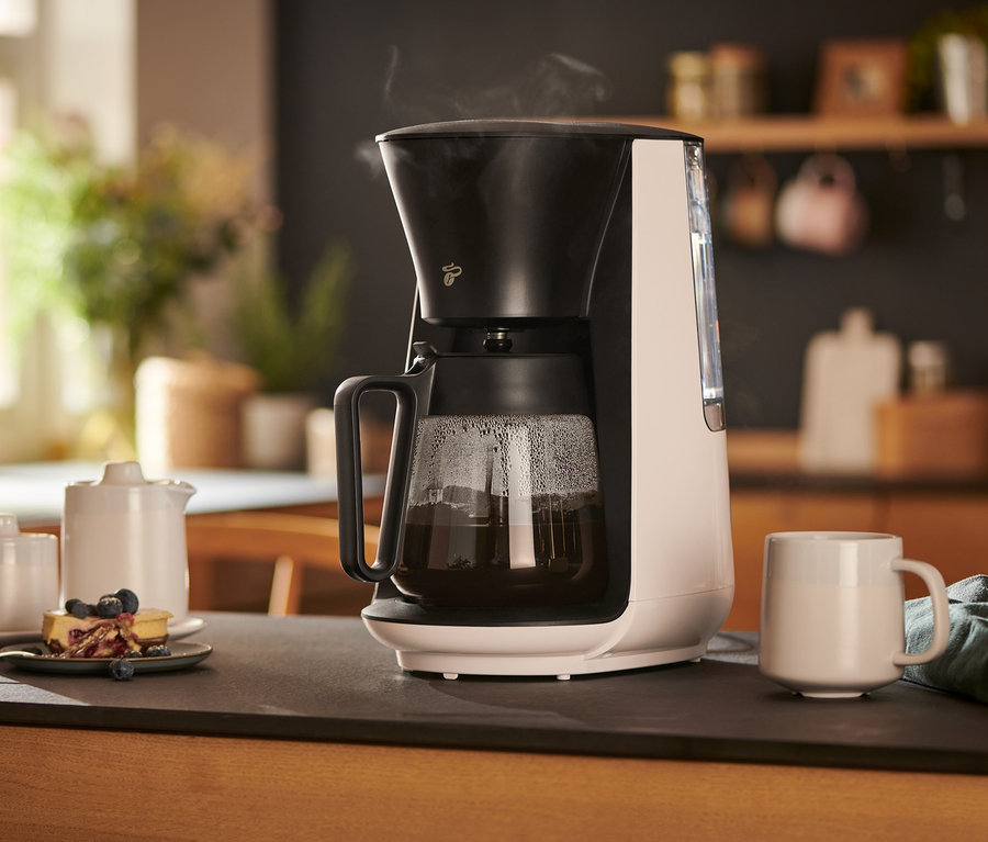 Egy fehér »Let's Brew« filteres kávéfőző egy asztalon áll egy csésze, egy tejeskancsó, egy cukortartó és egy tányér sütemény mellett.