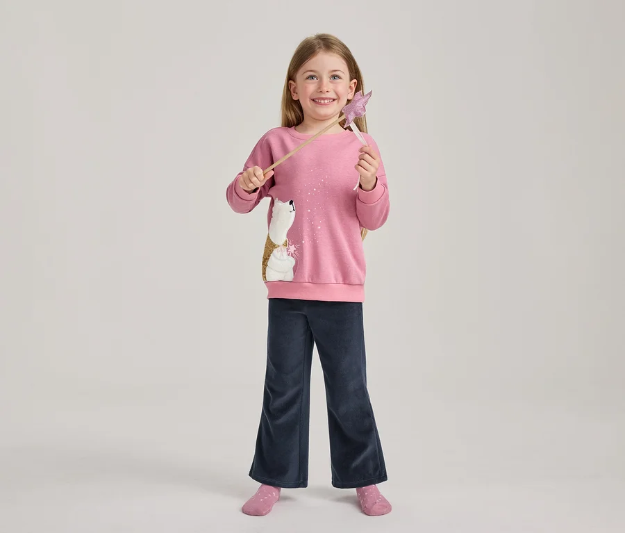 Mosolygó, hosszú hajú lány rózsaszín pulóverben, flitteres jegesmedve mintával, sötétkék bő szárú nadrágban és rózsaszín zokniban, rózsaszín csillagpálcát tartva.