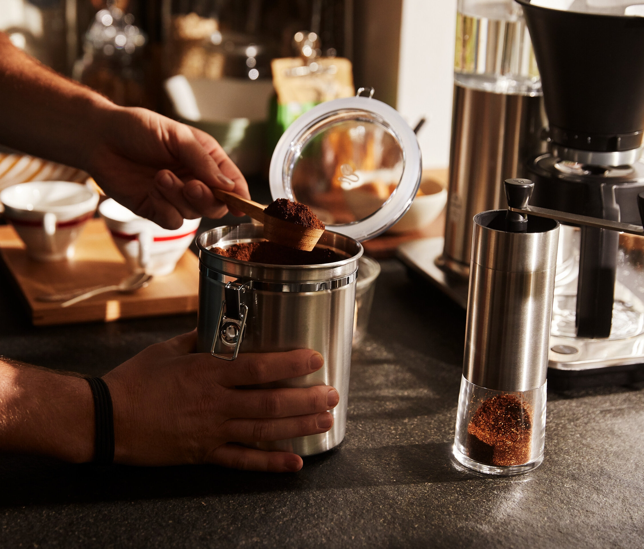 Férfi egy kanállal kávét merít egy fémdobozból. Mellette egy ezüst kézi kávédaráló áll.