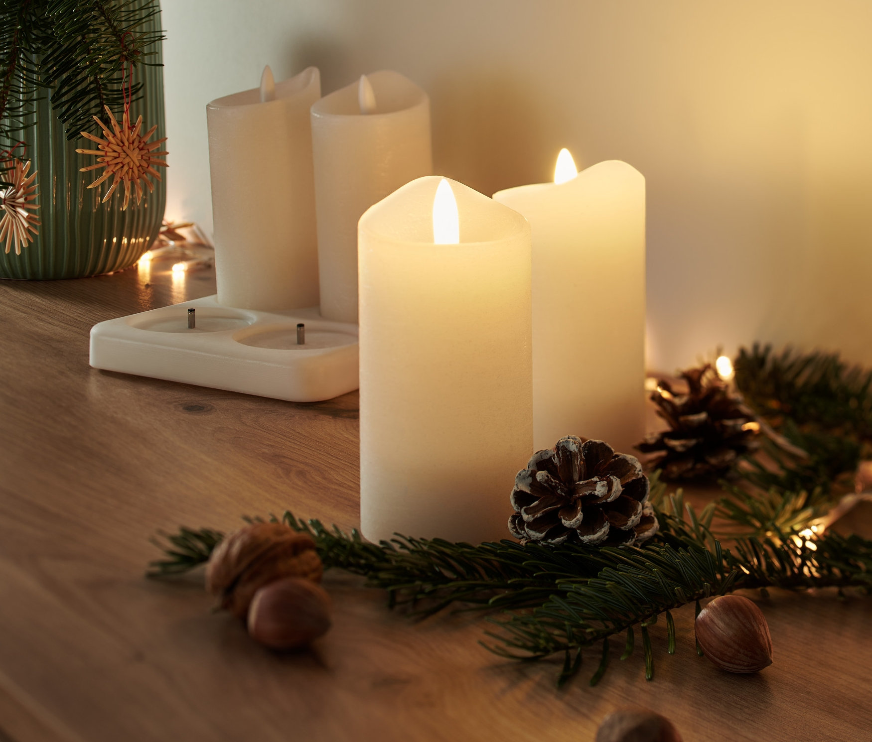 Négy LED-es valódi viaszgyertya akkumulátorral egy fa asztalon áll egy fenyőágakkal, tobozokkal és dióval teli váza mellett.