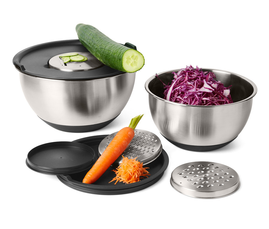 Rozsdamentes acél keverőtál készlet uborkával, vörös káposztával és sárgarépával egy reszelőn a háttérben.