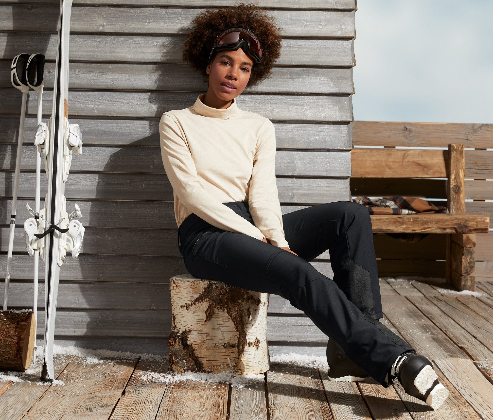 Nő ül egy fatönkön fekete softshell jet sínadrágban és síbakancsban.