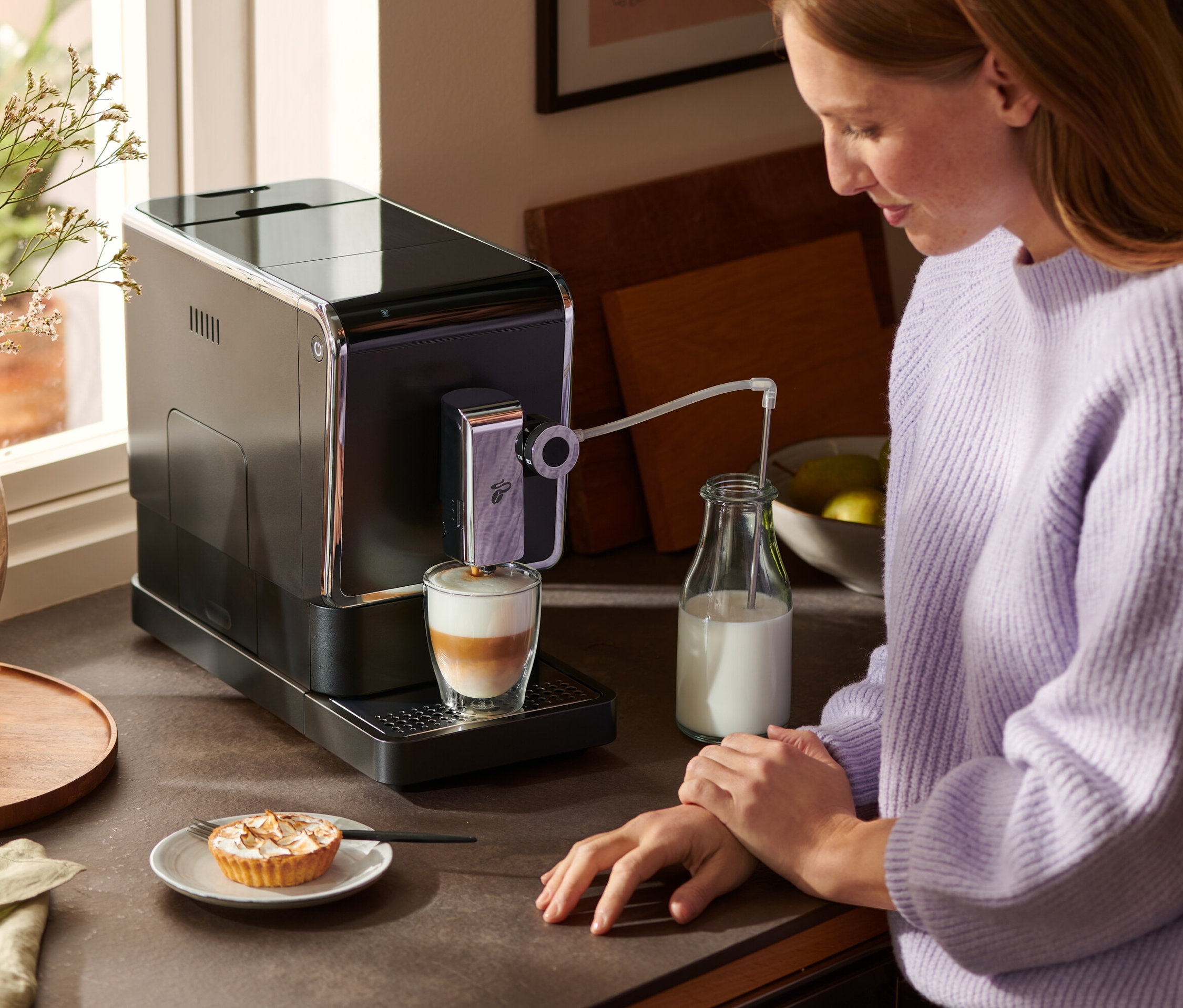 Egy nő a pultnál áll és egy antracit színű »Esperto Pro« Tchibo automata kávéfőzővel lattét készít. A kávéfőző mellett egy üveg tej és egy tányér sütemény áll.