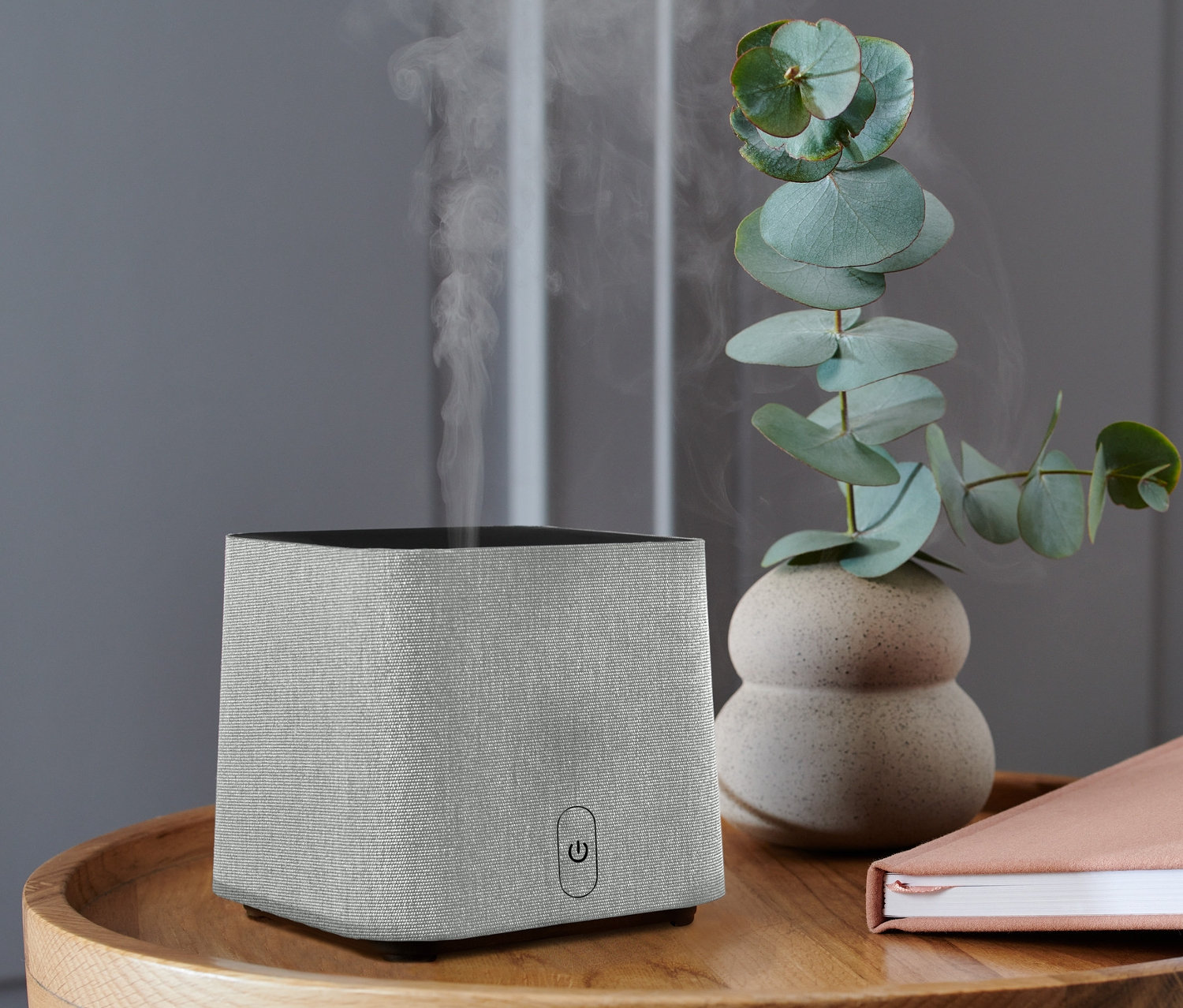Szürke aroma diffúzor egy fa asztalon, könyvvel és eukaliptusz vázával.