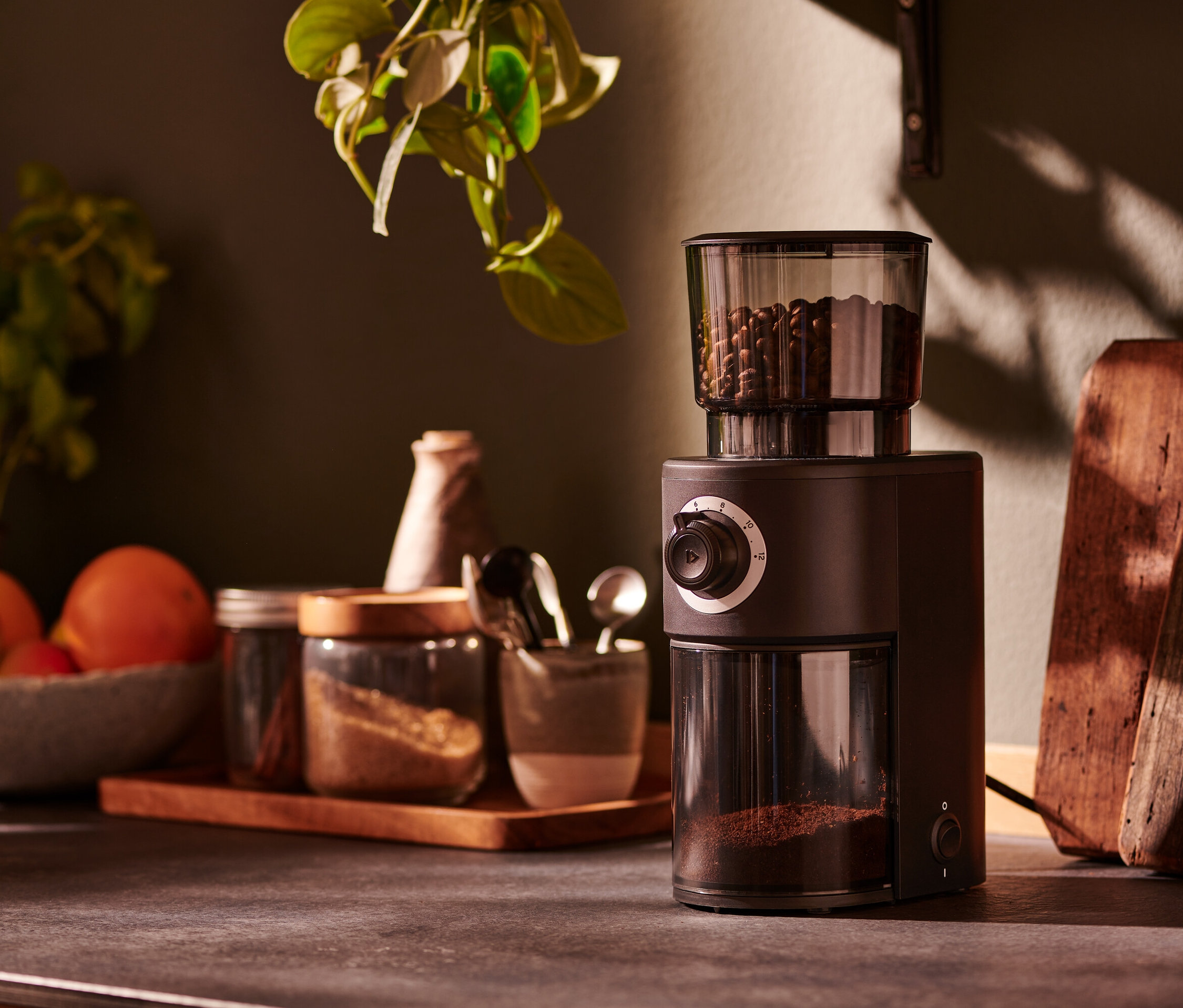 Egy barna, elektromos kávédaráló áll egy konyhapulton fűszerekkel teli üvegek mellett.