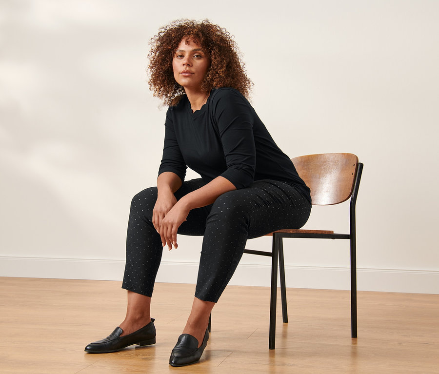 Egy nő ül egy széken, fekete 3/4-es ujjú felsőt és pöttyös sztreccsnadrágot visel.
