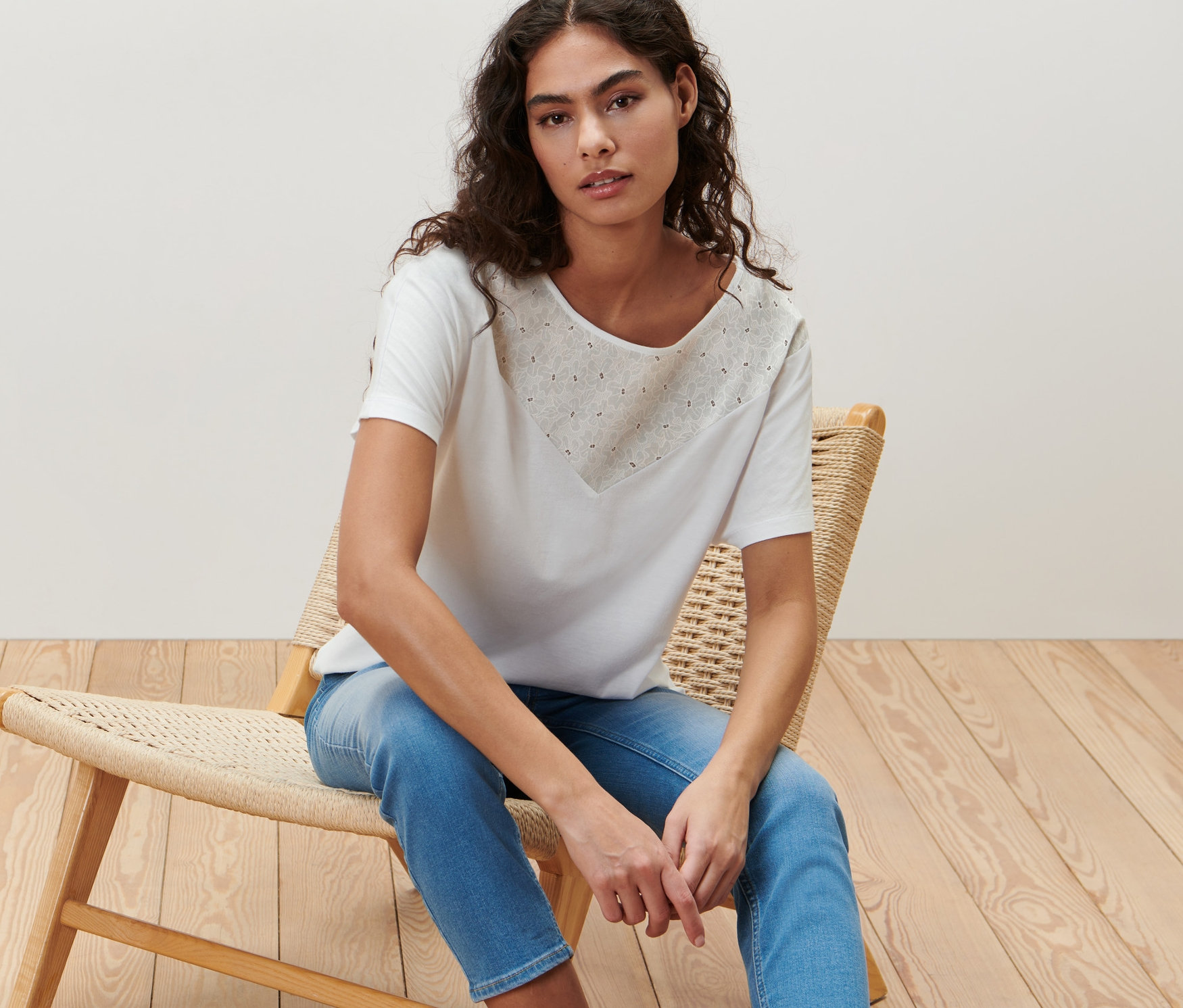 Nő ül egy fonott széken, és fehér, hímzett pólóblúzt visel.