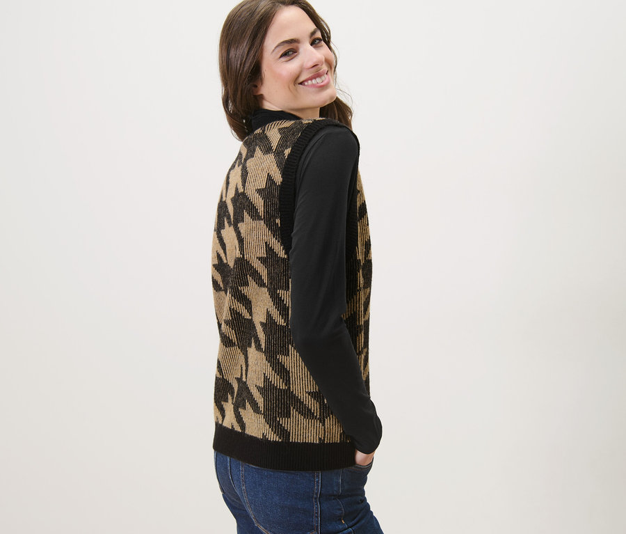 Egy nő mosolyog a válla fölött, és tyúklábmintás kötött mellényt visel.