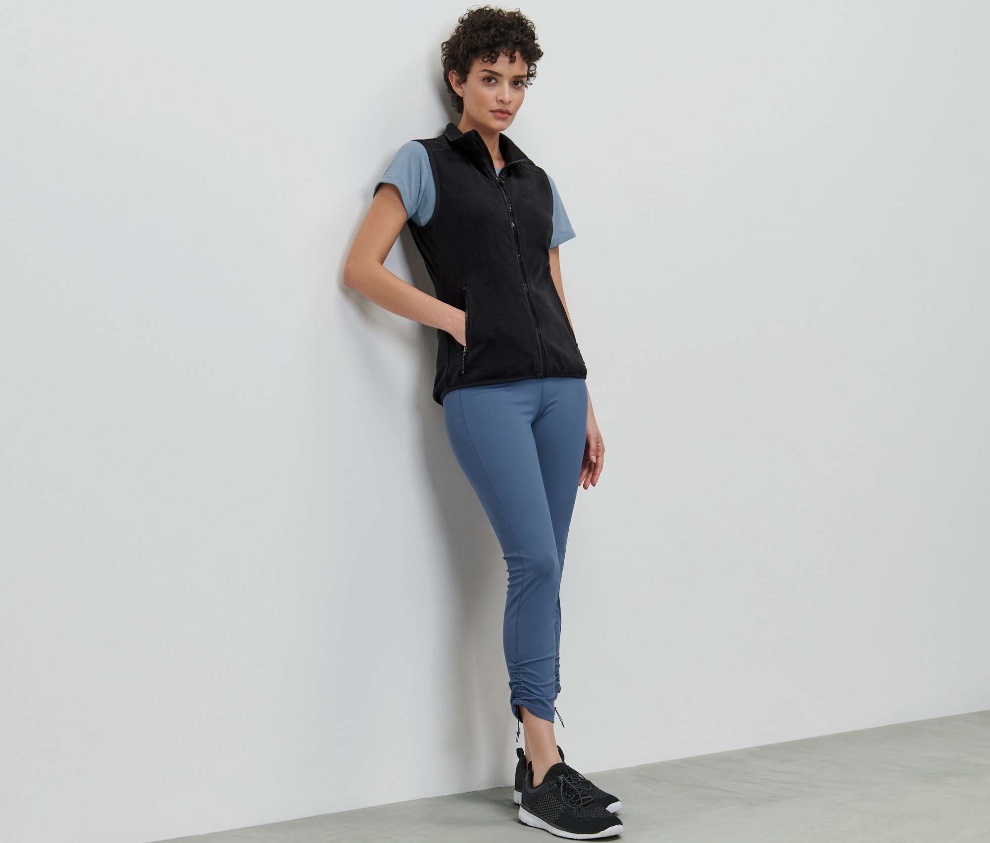 Egy nő a falnak dőlve fekete polármellényt, pólót és leggingst visel.