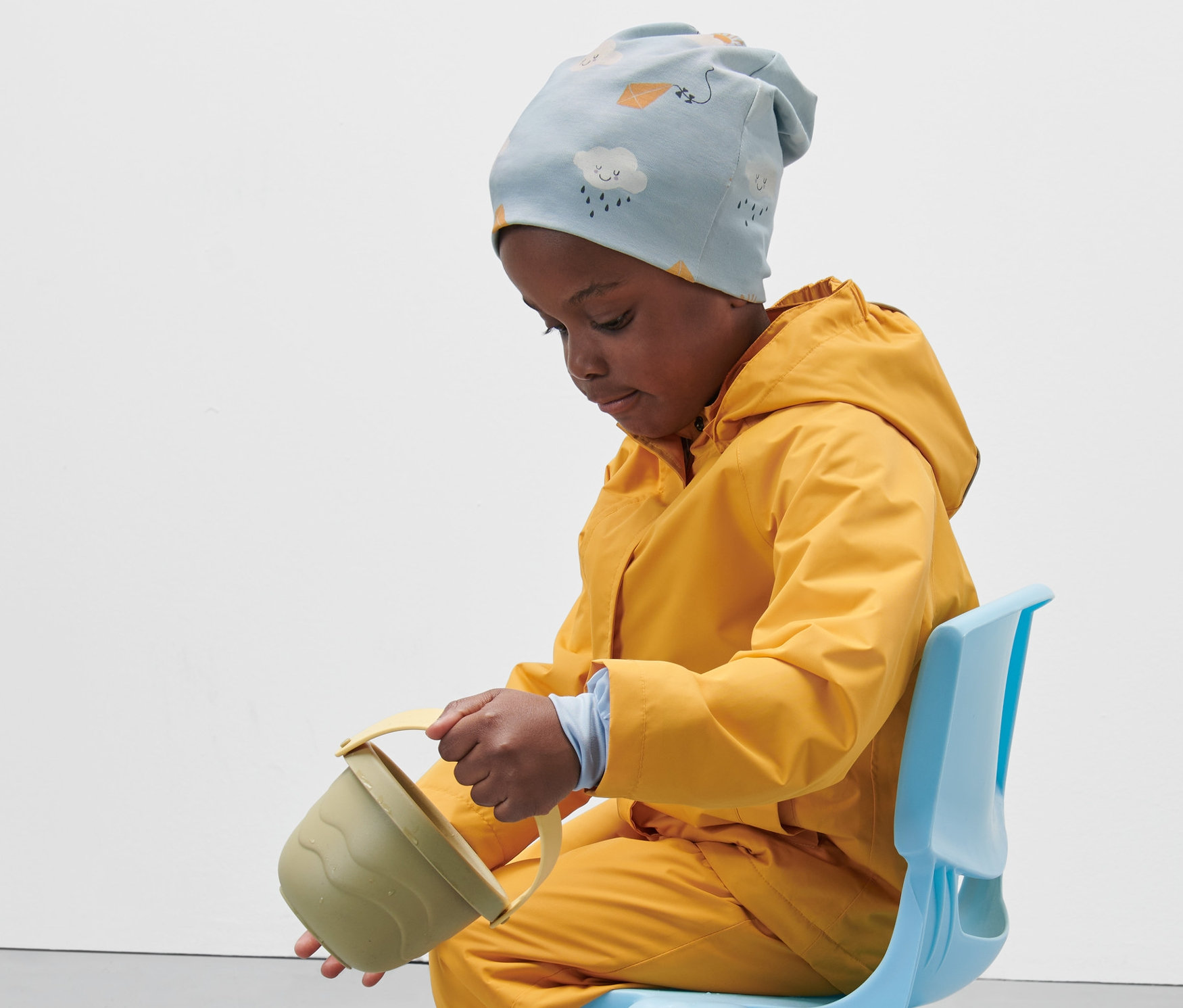 Egy gyerek ül egy kék széken, sárga esőkabátot és világoskék gyerek jersey sapkát visel, és vizet önt egy játékvödörből.