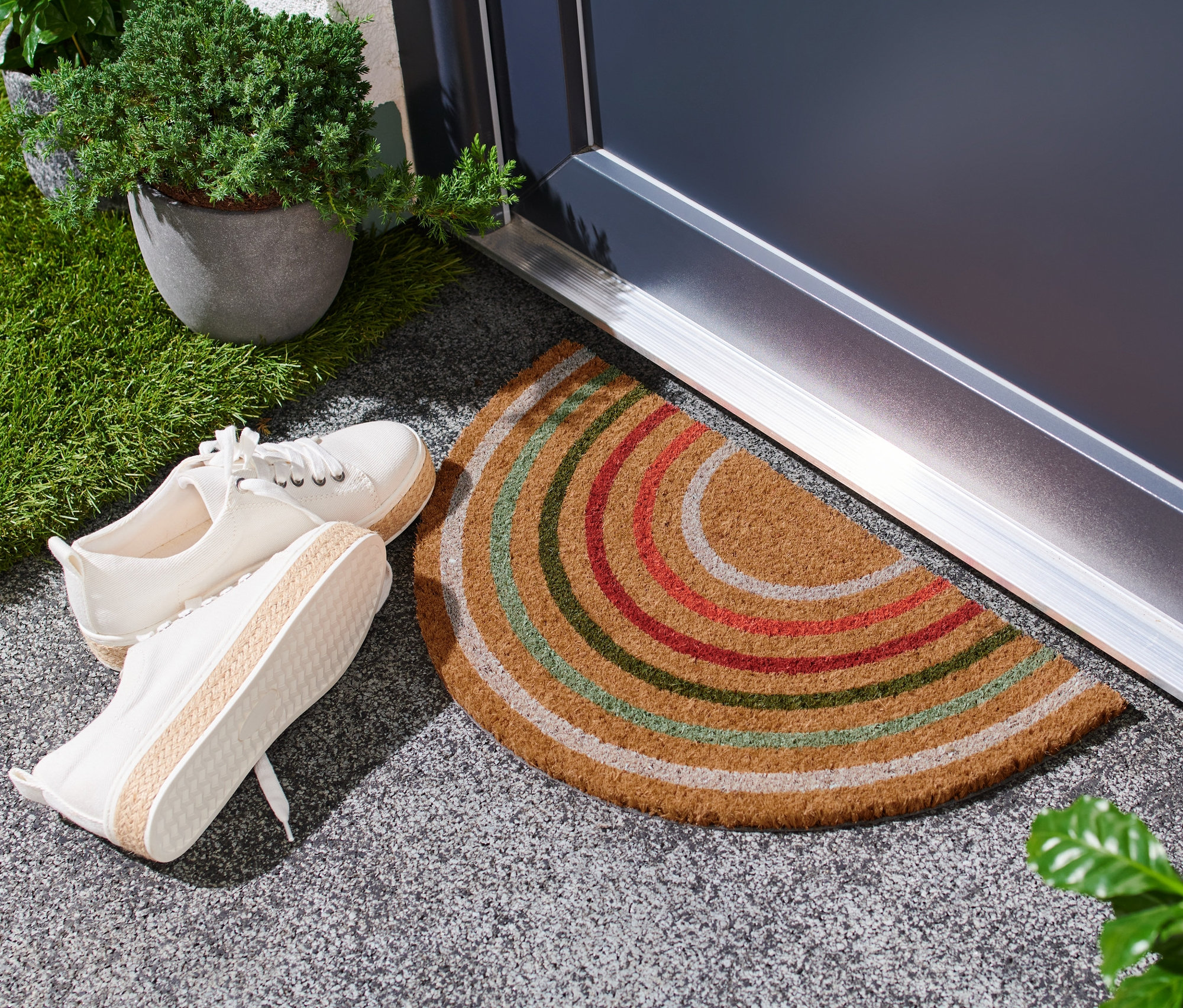 Egy ajtó előtt egy színes csíkos kókusz lábtörlő fekszik, mellette egy pár fehér cipő áll.