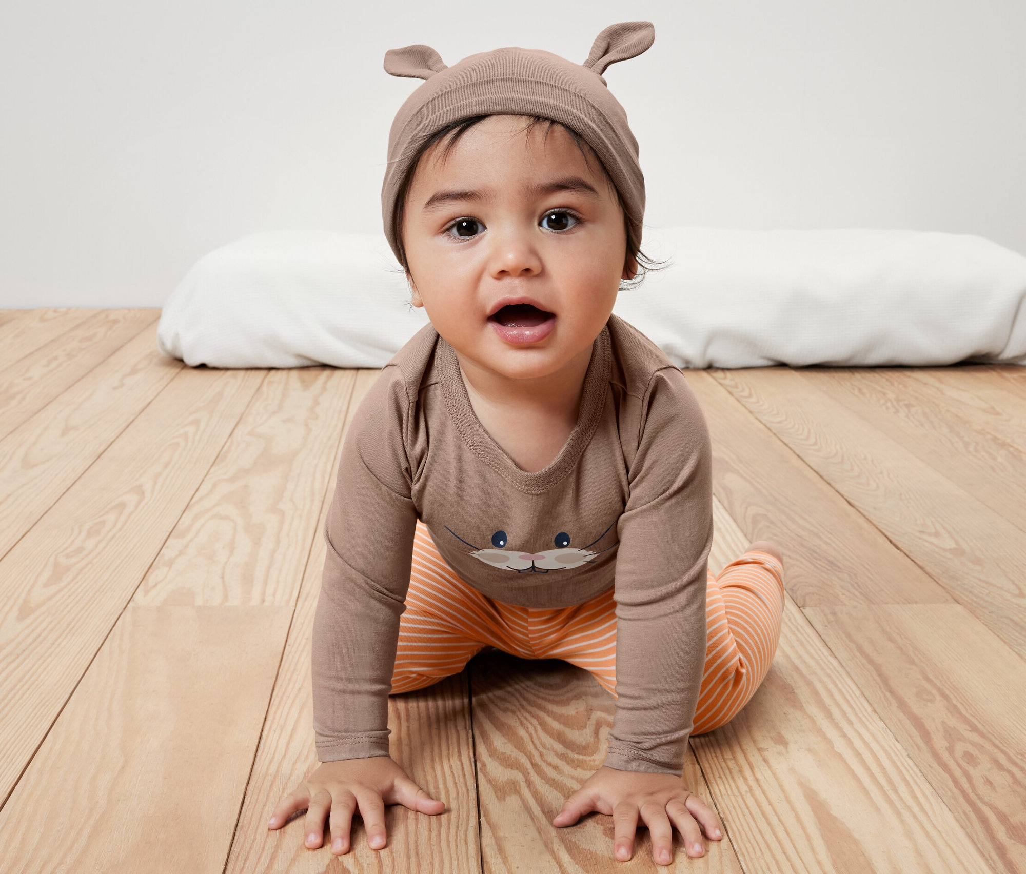 Kisgyerek háromrészes együttes, nyuszis: A baba barna, mintás bodyban és narancssárga csíkos nadrágban mászik a fapadlón. Barna füles sapka.