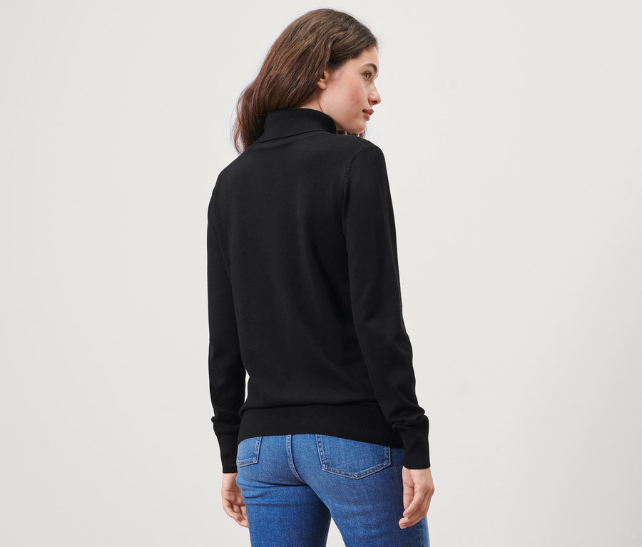 Egy nő háttal áll a kamerának, fekete, finom kötésű garbót és farmert visel.