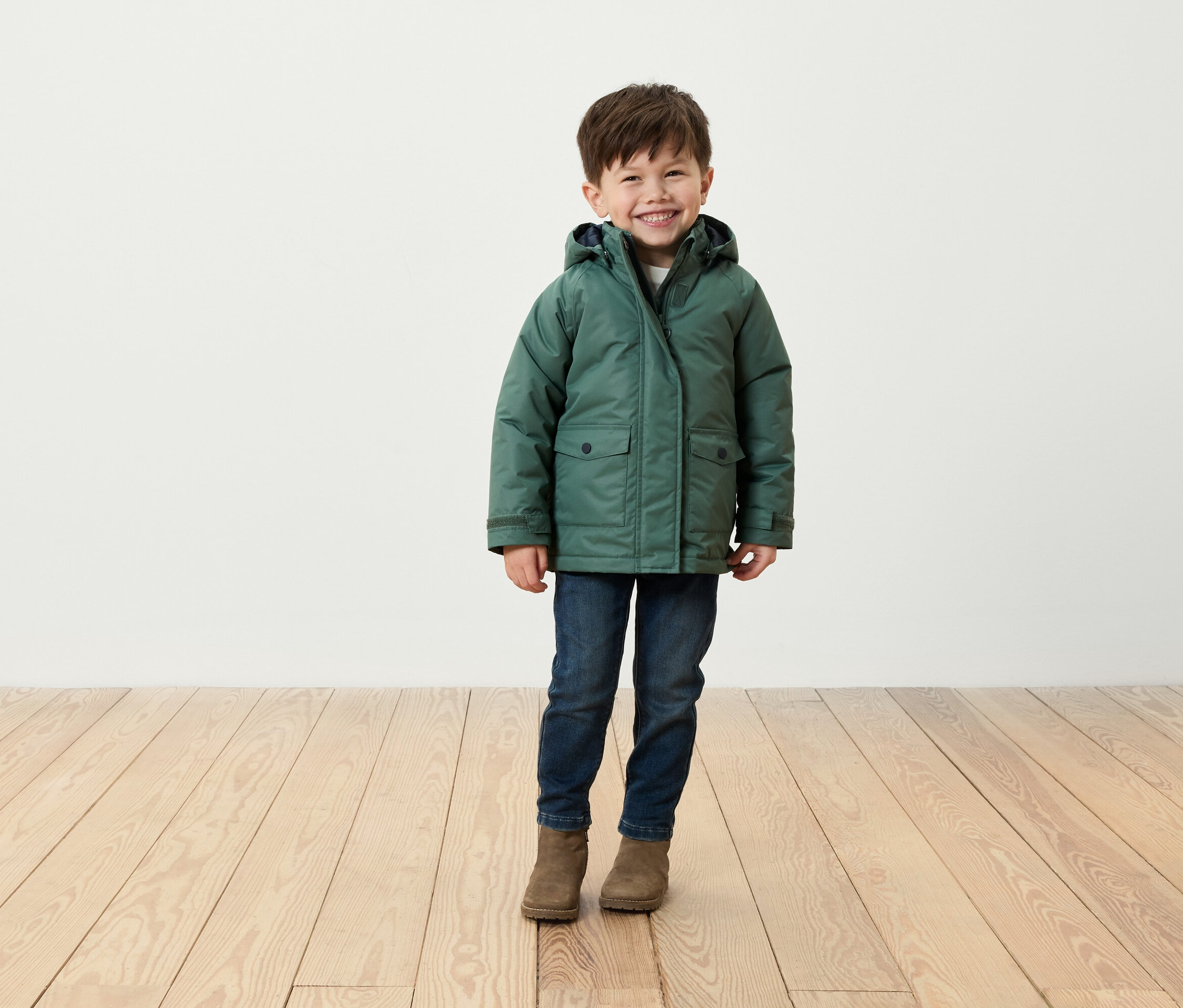 Egy mosolygó fiú áll egy fapadlón, zöld kabátot, farmert és barna csizmát visel.