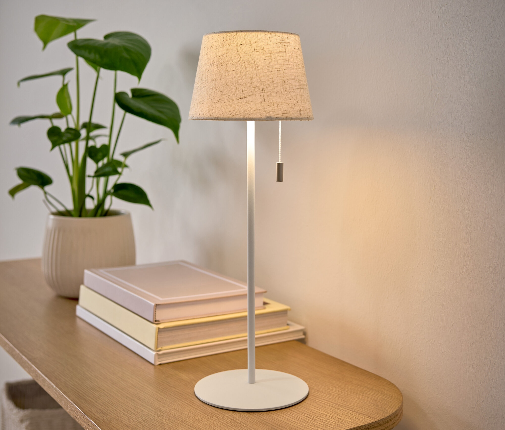 Egy bézs lámpabúrás asztali lámpa áll egy faasztalon egy cserepes növény és egy könyvkupac mellett.