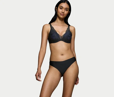 Sötét hajú nő fekete Triumph Body Makeup Illusion WP csipkemelltartót visel kék béléssel és fekete alsóneműt, fehér háttér előtt állva.