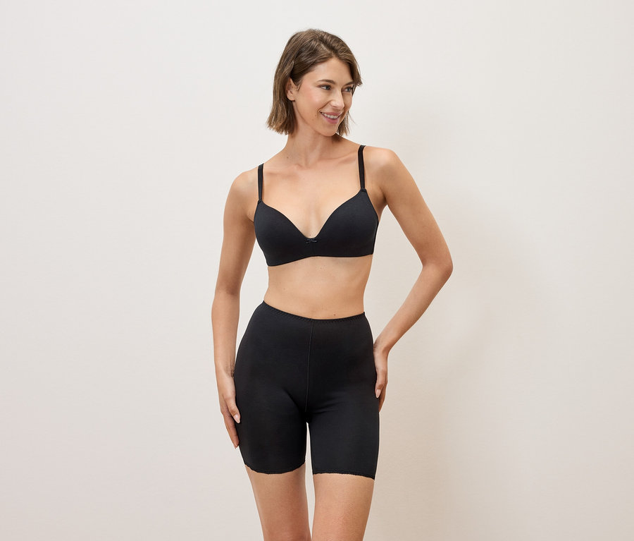 Nő fekete melltartóban és fekete rövidnadrágban pózol semleges háttér előtt.