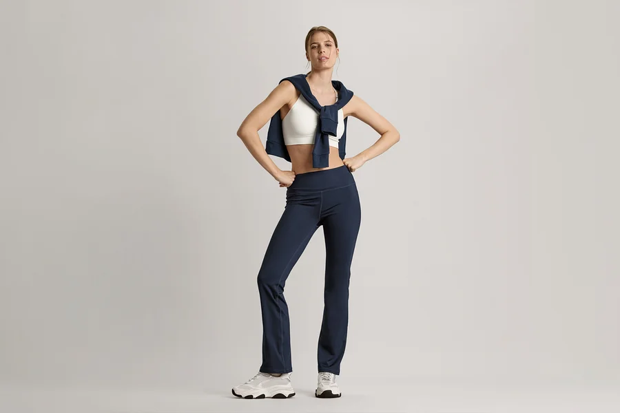 Frau posiert mit Sweatjacke in Navy um die Schultern, weißem Seamless-Sport-Bustier und marineblauer Bodyforming-Sporthose.