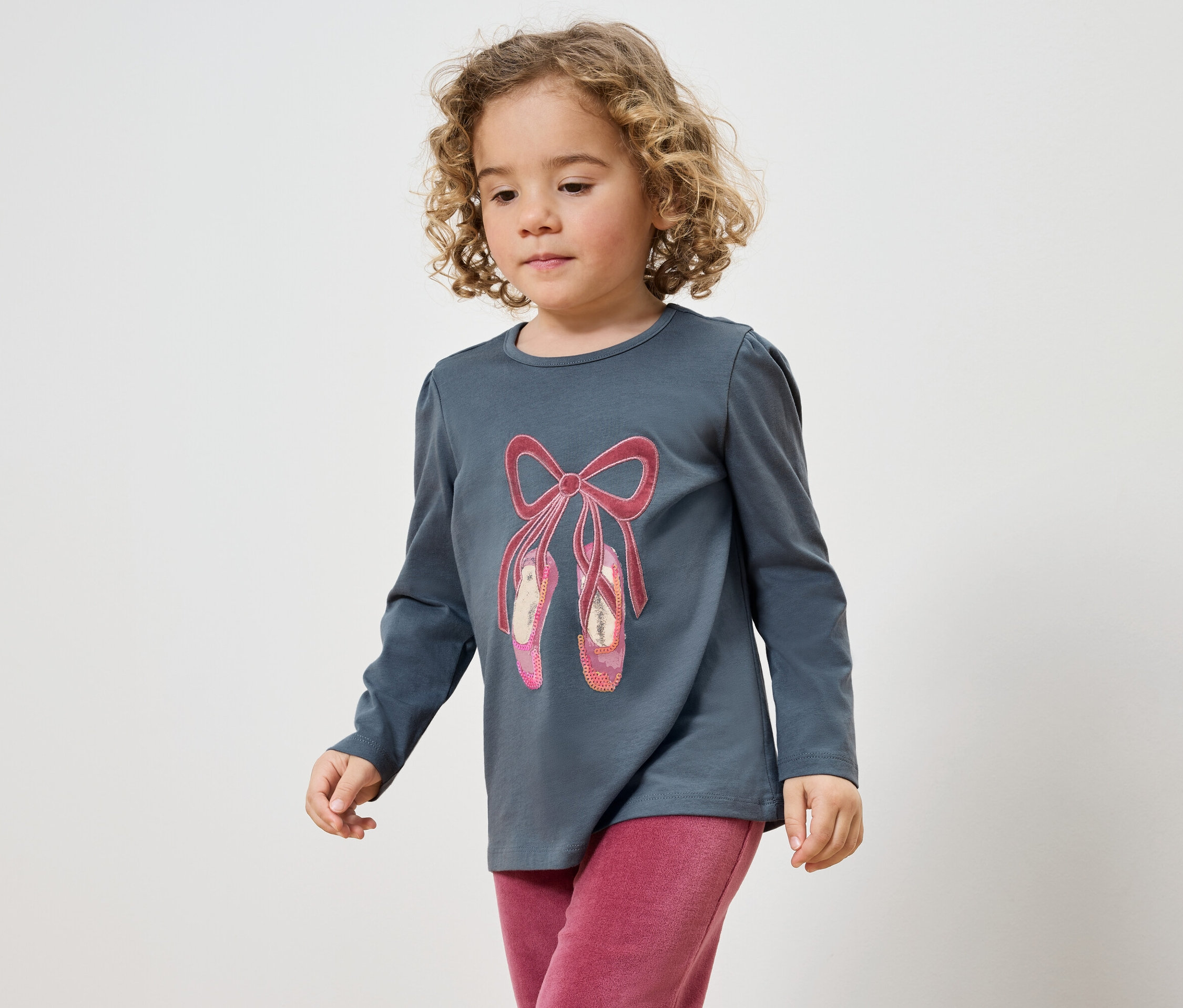 Göndör hajú kislány szürke pólóban, balettcipő mintával és rózsaszín nadrágban.