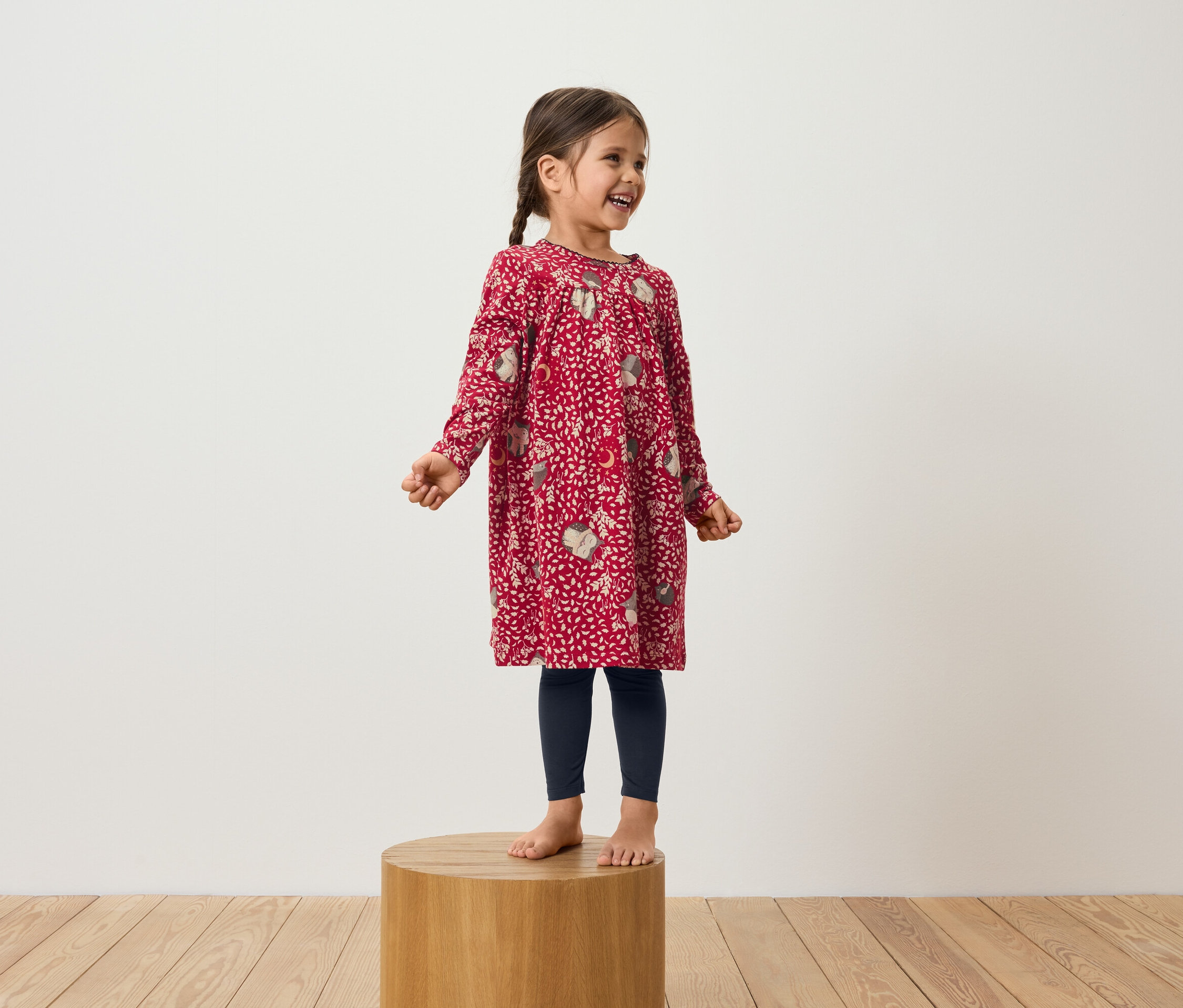 Egy mosolygó kislány áll egy fa emelvényen, piros, hosszú ujjú ruhát és sötétkék leggingset visel.