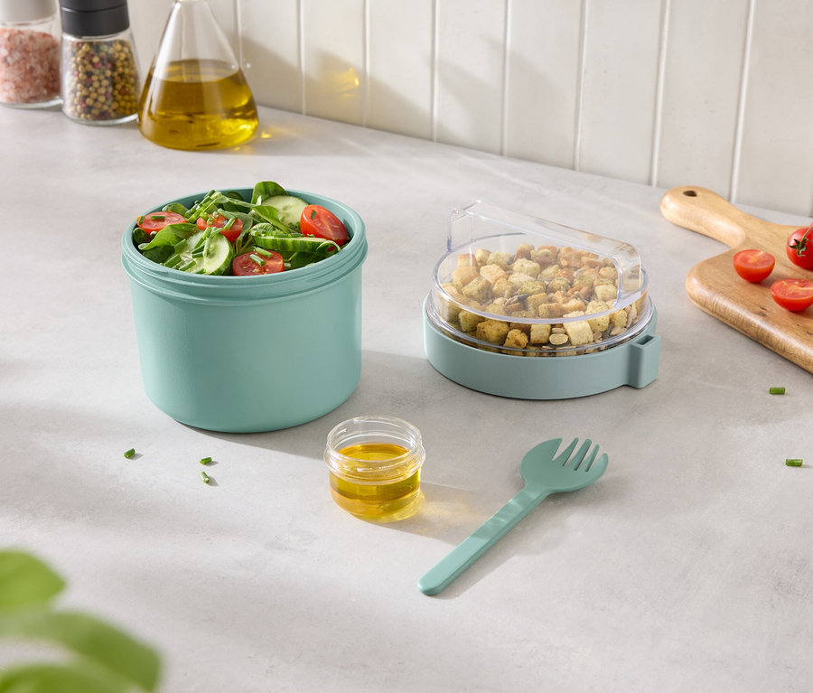 Vegyes saláta türkizkék edényben, krutonnal átlátszó fedéllel ellátott edényben, valamint öntettel és villával mellette.