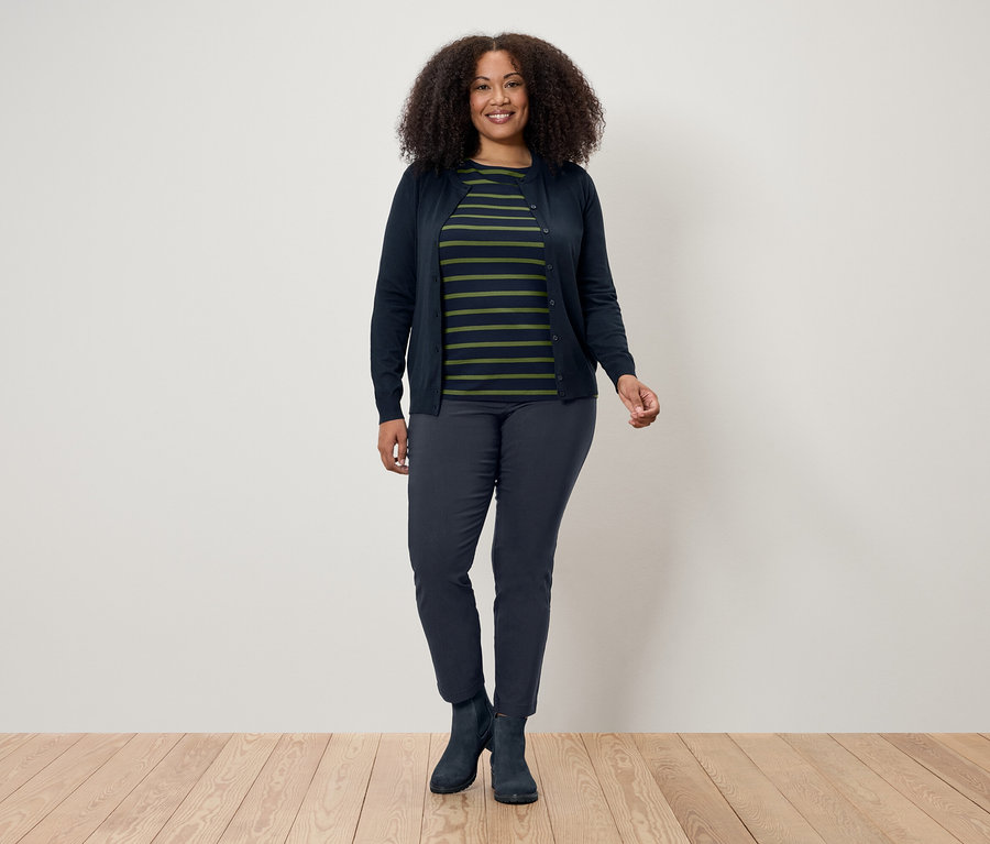 Egy göndör hajú nő áll egy fapadlón egy fehér fal előtt. Sötétkék kardigánt, csíkos pólót, sötétkék nadrágot és csizmát visel.